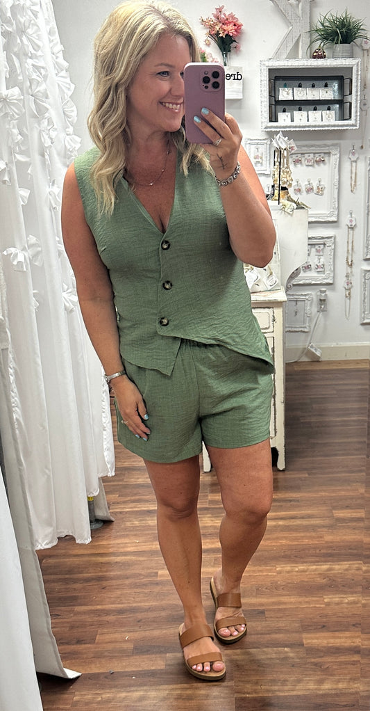 Woman taking a mirror selfie wearing a green outfit in a room with white curtains and a shelf.