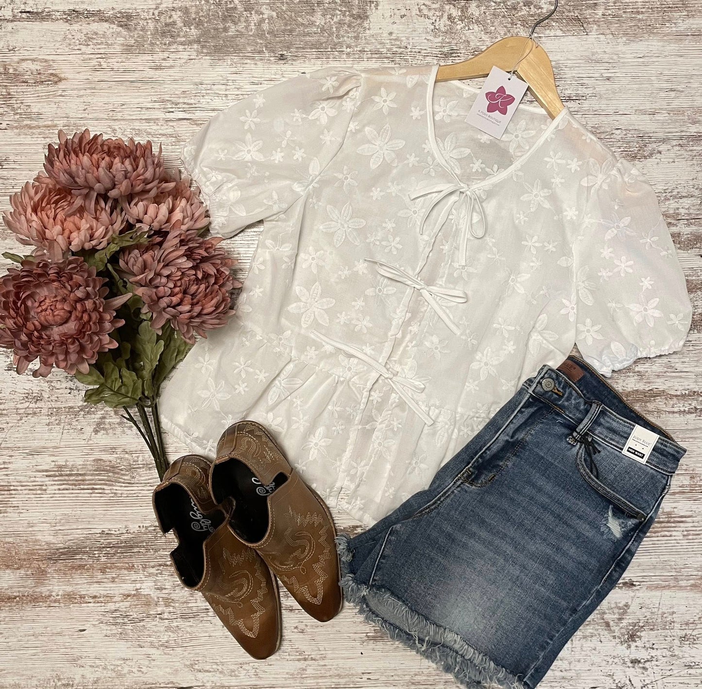 White lace top with a hanger, brown shoes, and blue jeans on a wooden surface with flowers.