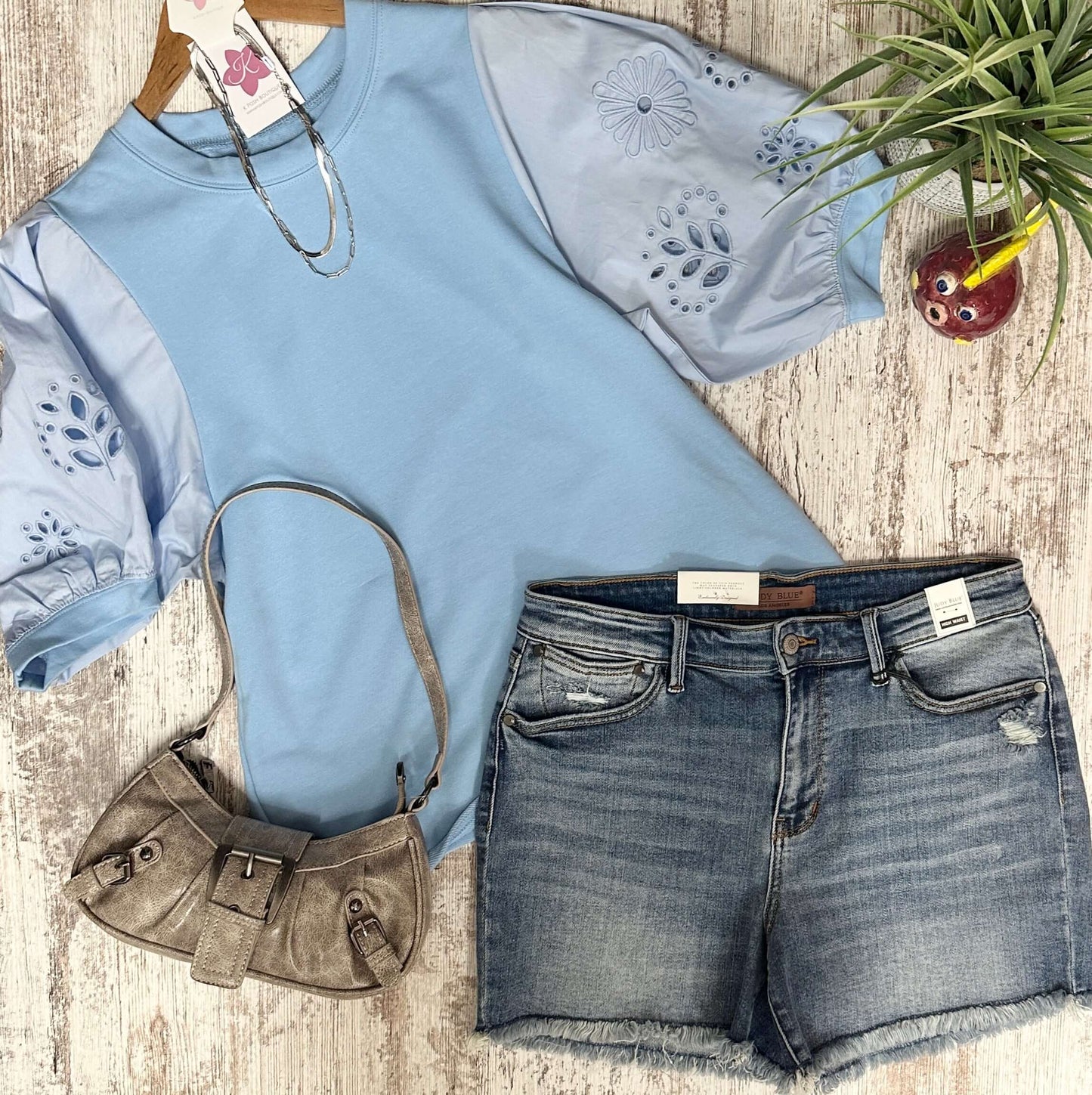Light blue shirt with floral cut-out sleeves, denim shorts, and brown handbag on a wooden surface.
