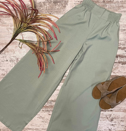 Green pants on a wooden surface with a plant and sandals.