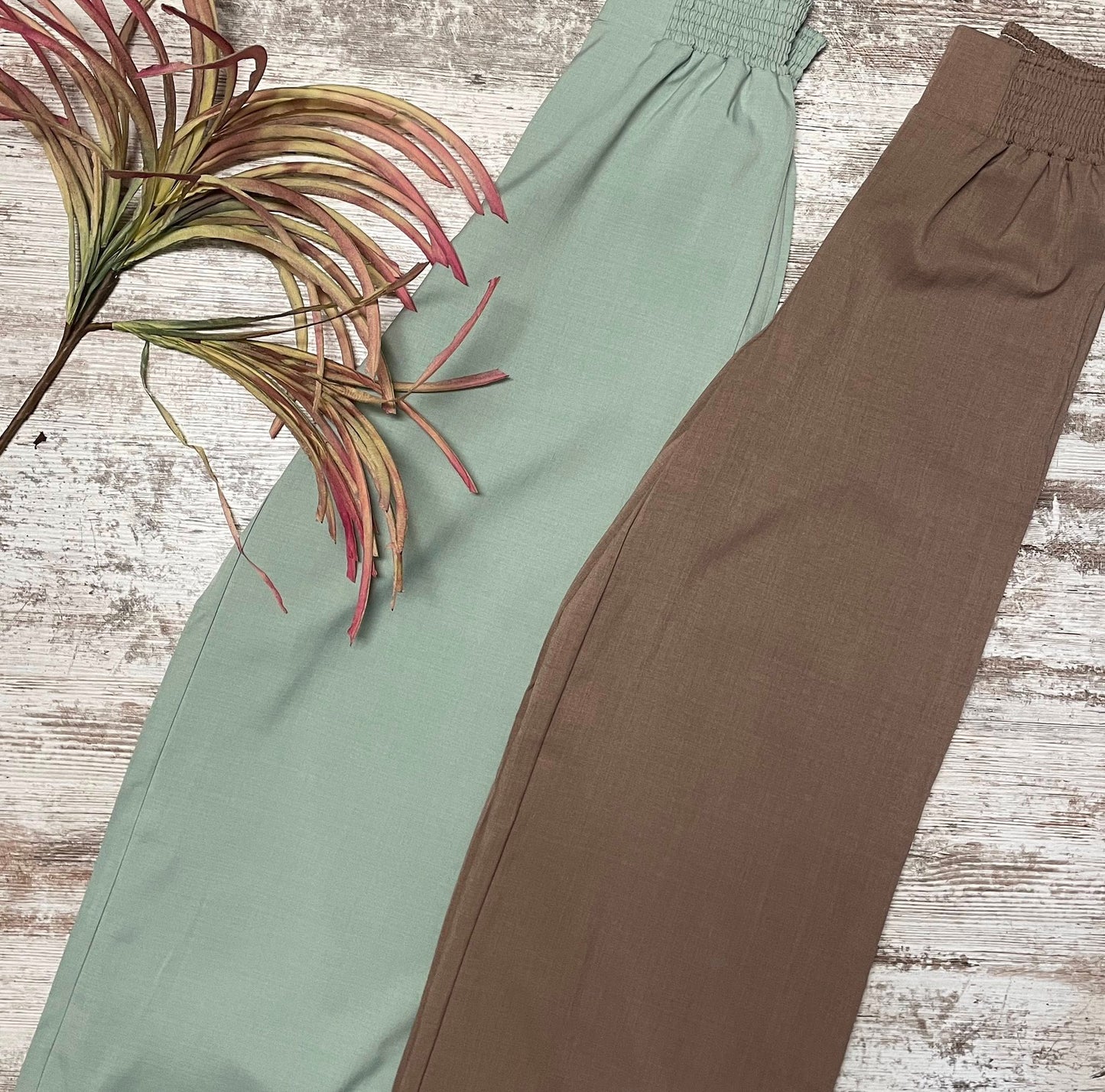 Two pairs of pants, one green and one brown, on a wooden surface with a plant leaf.