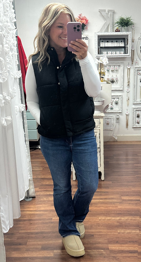 Person taking a mirror selfie wearing a black vest, white shirt, blue jeans, and beige shoes in a store.