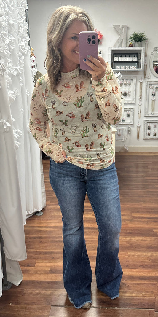Sheer mesh top featuring a playful cactus and boots print in earthy tones, styled on a model with high-waisted denim. The lightweight fabric and western-inspired design make it perfect for layering or wearing solo for a bold statement look.
