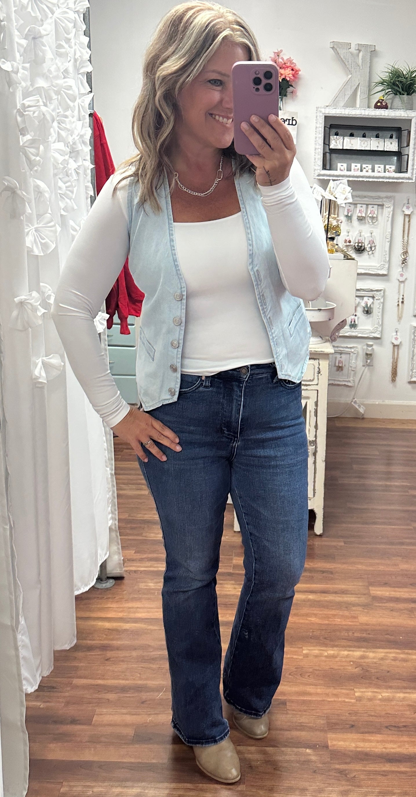 Woman taking a mirror selfie wearing a light blue vest, white shirt, and dark jeans.