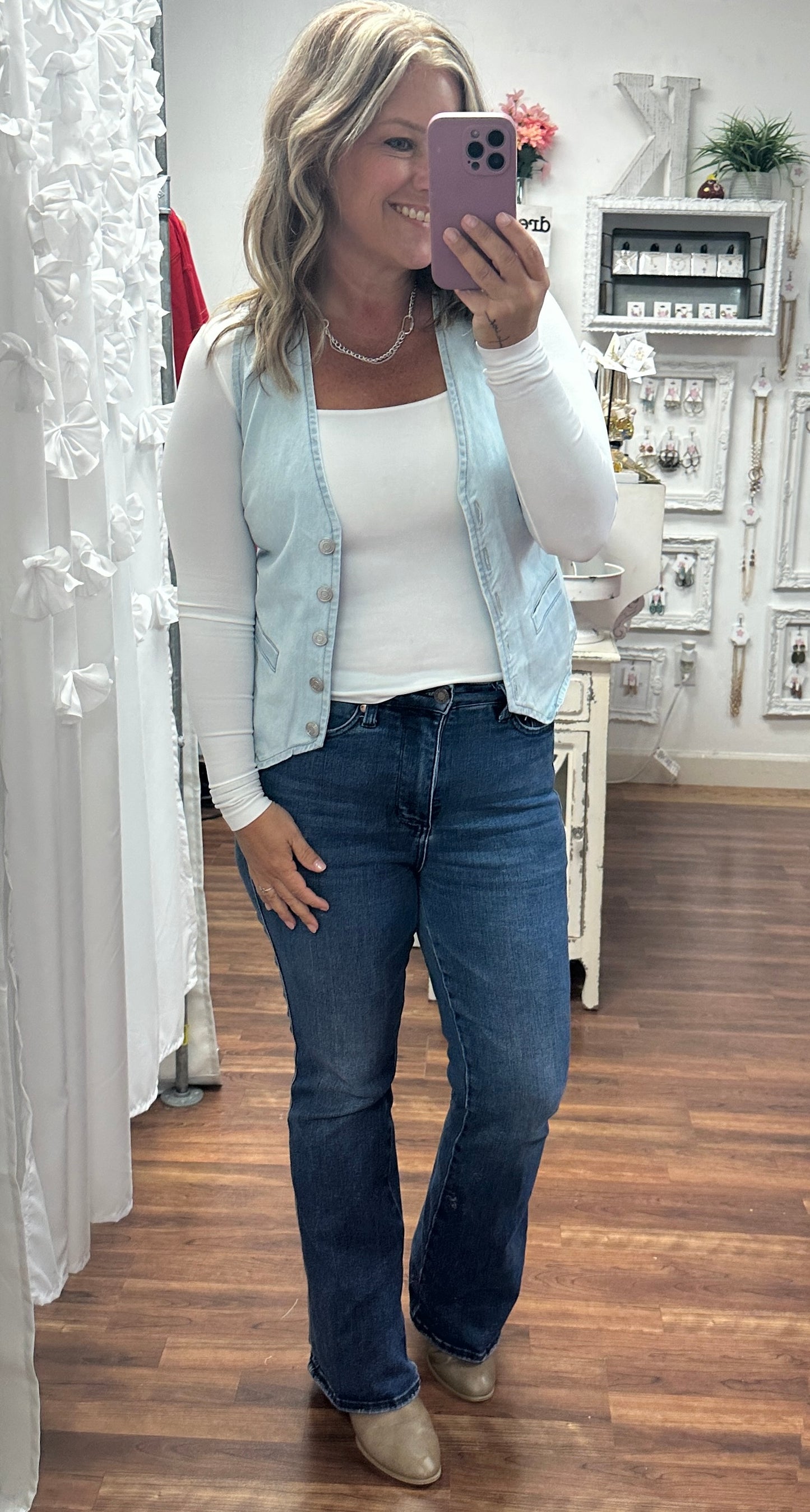 Woman taking a mirror selfie wearing a light blue denim vest, white long-sleeve top, and blue jeans.