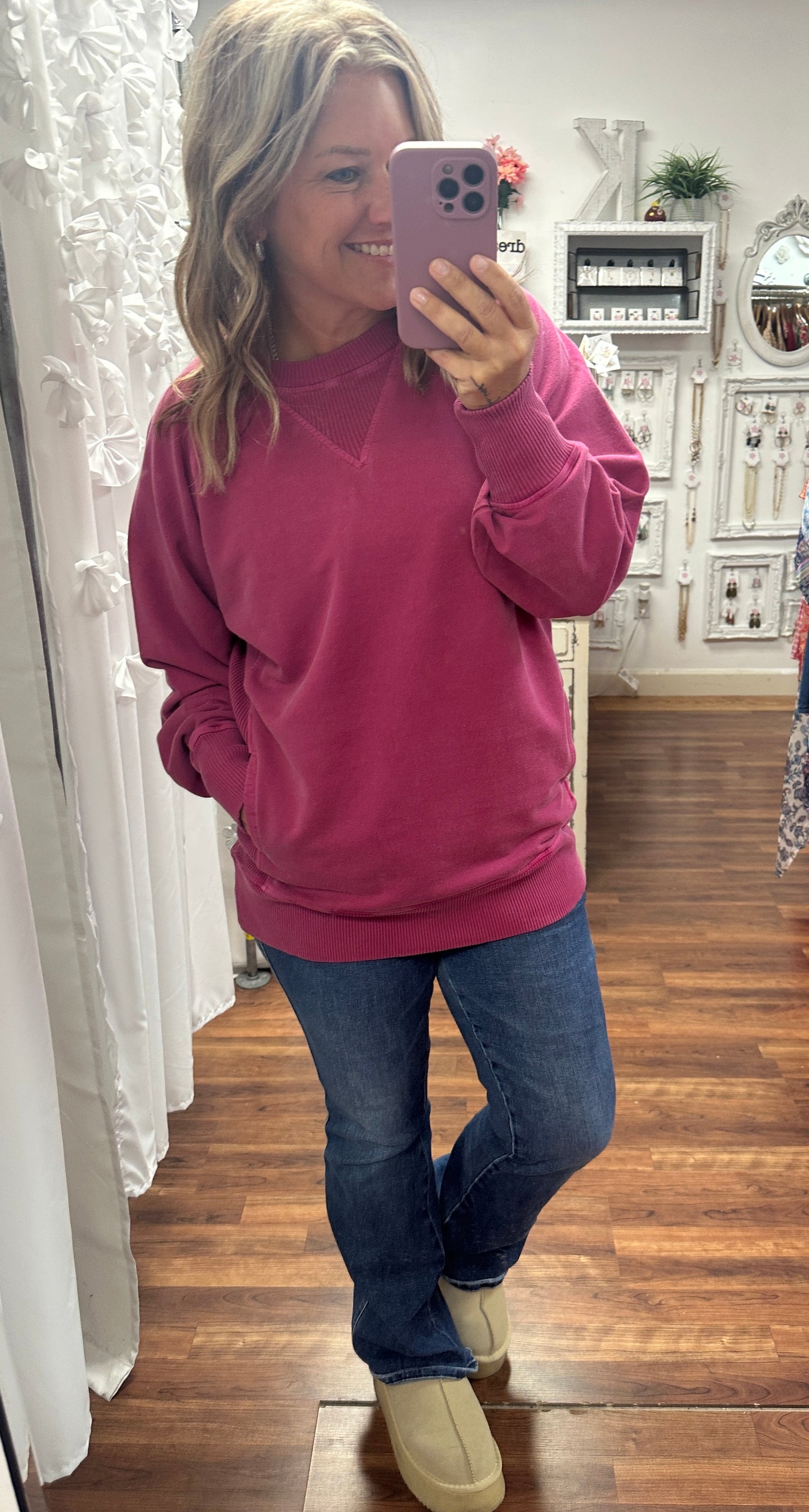 Woman taking a mirror selfie wearing a pink sweatshirt and blue jeans in a room with wooden flooring.