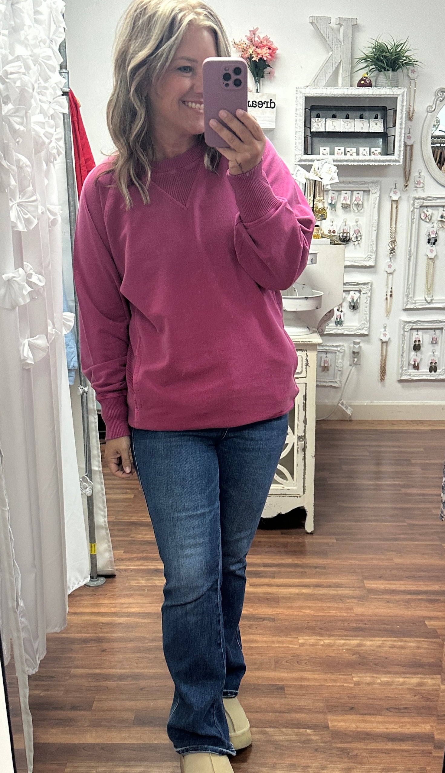 Person taking a mirror selfie wearing a pink sweatshirt and blue jeans in a store.
