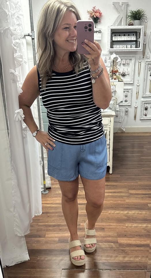 Woman taking a mirror selfie wearing a black and white striped tank top and blue shorts.