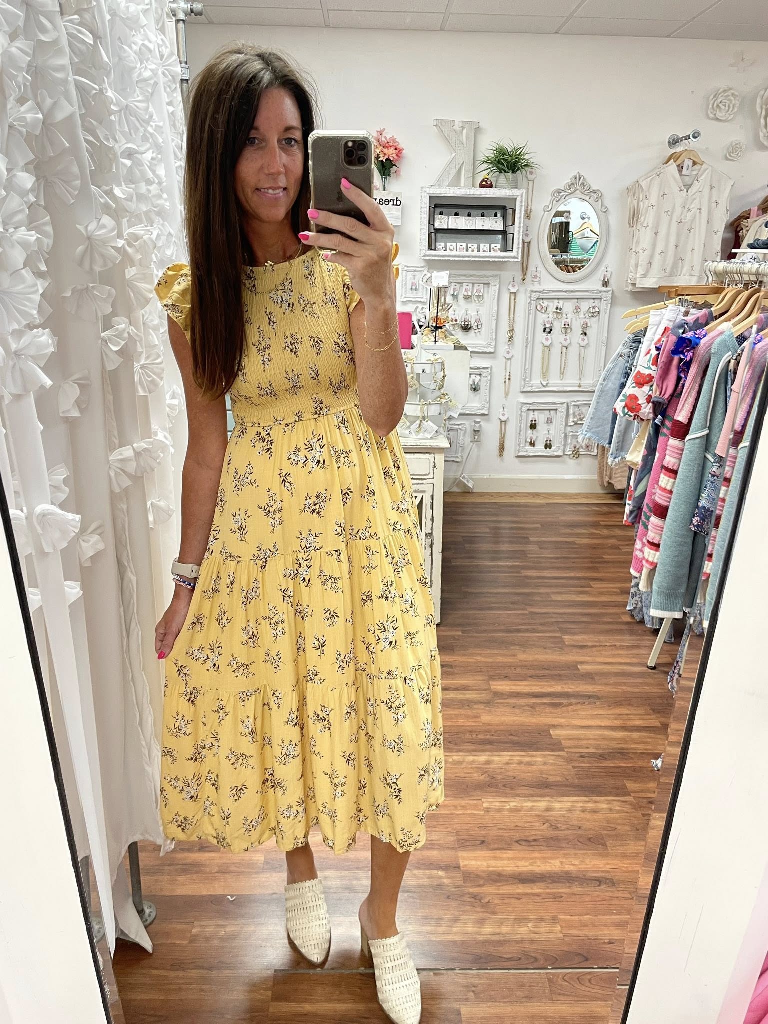 Woman taking a mirror selfie wearing a yellow floral dress in a clothing store.