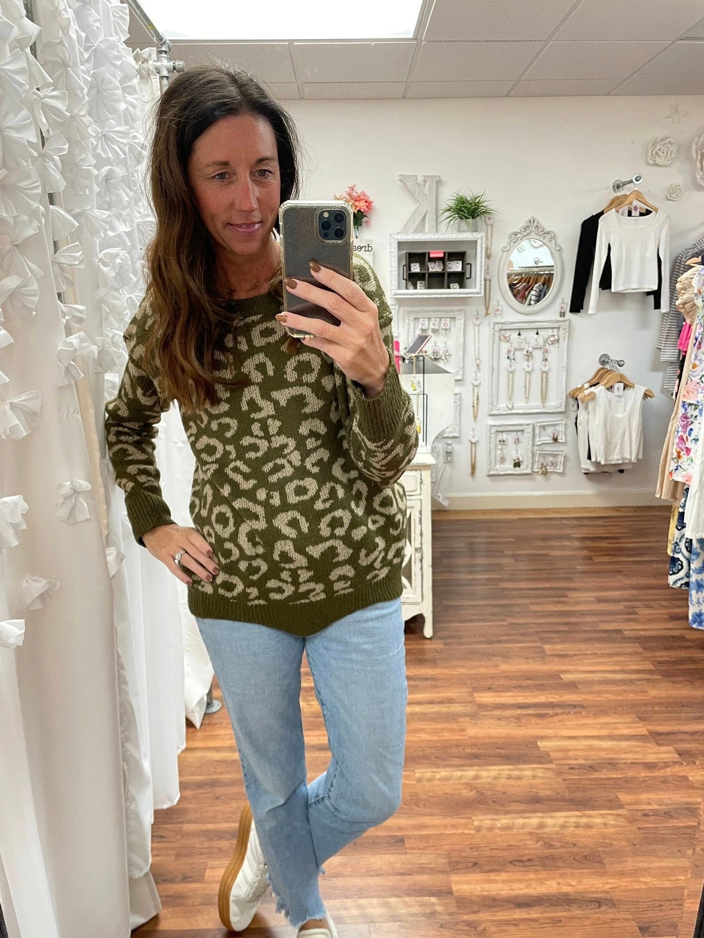 Woman taking a mirror selfie wearing a green leopard print sweater and light blue jeans in a store.