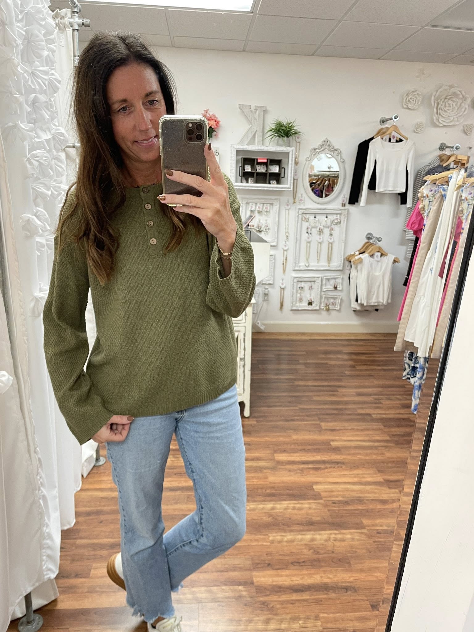 Woman taking a mirror selfie wearing an olive green sweater and light blue jeans in a clothing store.