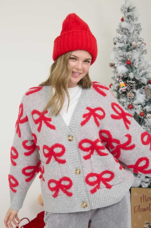 Person wearing a gray cardigan with red bows and a red beanie in front of a Christmas tree.